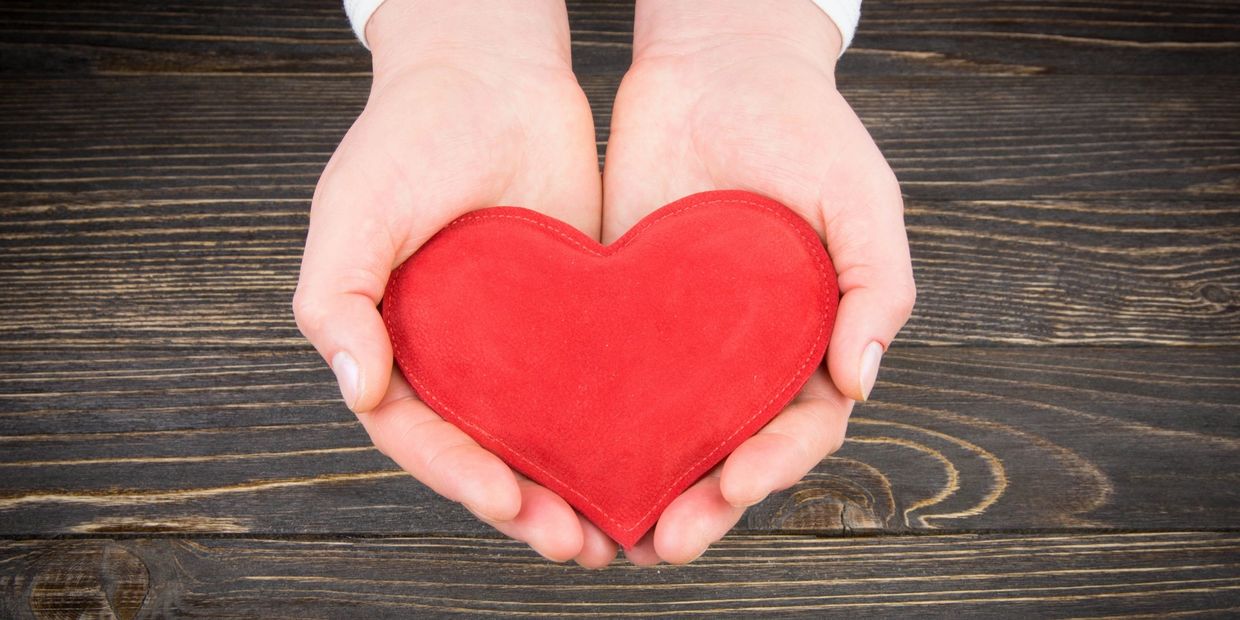 Hands gently holding a red heart on a wooden surface.