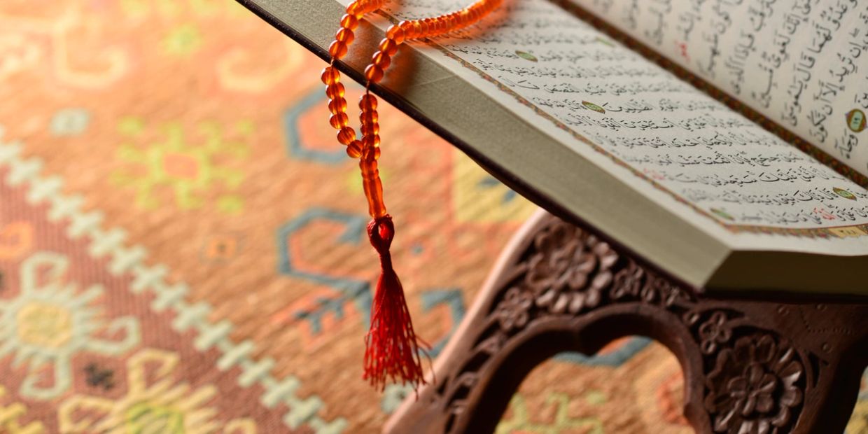An open Quran with prayer beads on a wooden stand over a patterned carpet.
