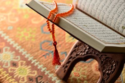 Open Quran on a wooden stand with prayer beads on a patterned carpet.